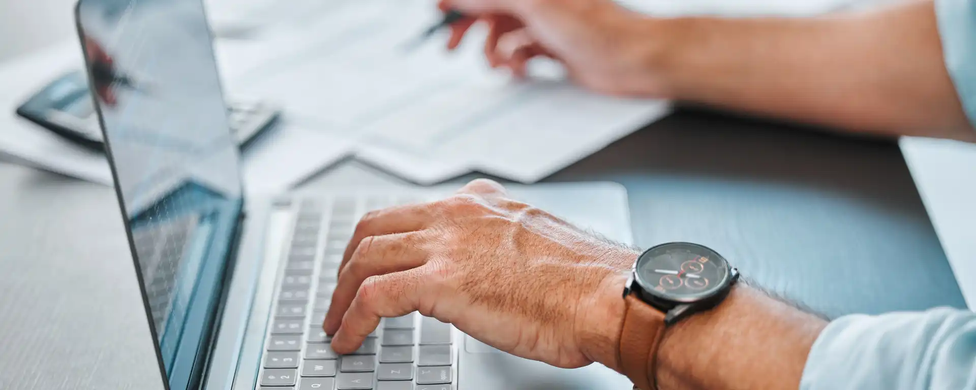 Eine Hand, die auf der Tastatur eines Laptops liegt, im Hintergrund eine andere Hand, die etwas notiert 