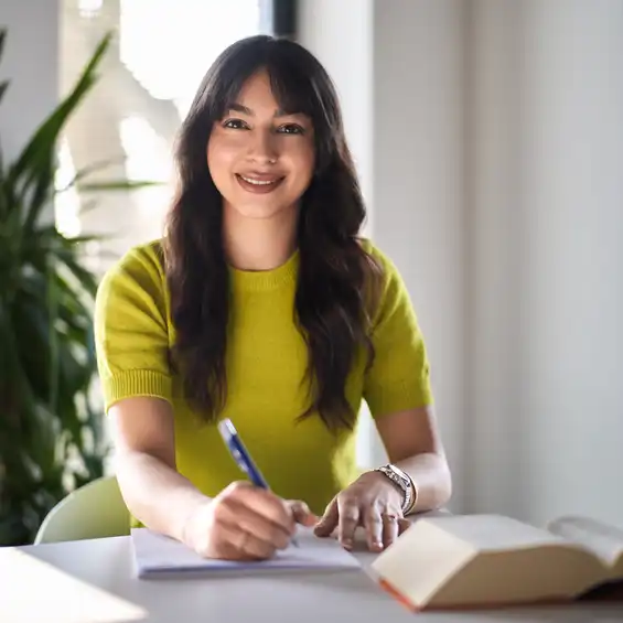 Eine Frau sitzt am Tisch und schreibt etwas auf, vor ihr liegt ein dickes Buch